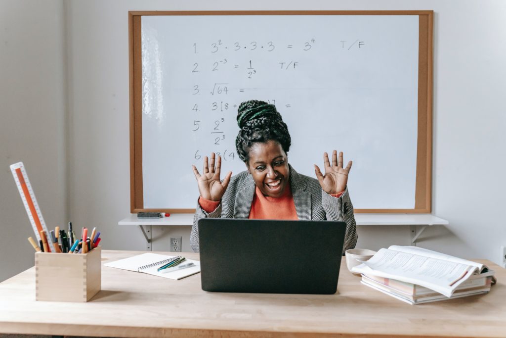 teacher smiling and waving at laptop screen in front of them while seated in a classroom.