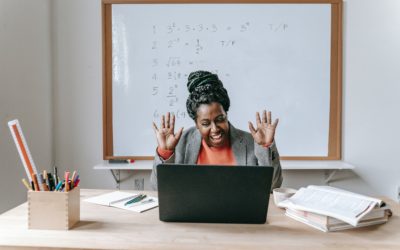 teacher smiling and waving at laptop screen in front of them while seated in a classroom.